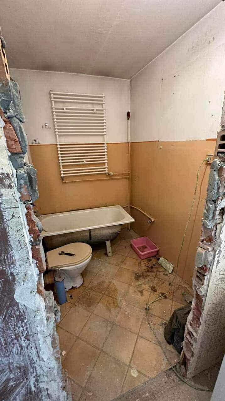 Łazienka w trakcie demontażu: stara wanna, toaleta, grzejnik na ścianie, zerwane płytki, gruz. Widok przed remontem, zniszczone wnętrze.