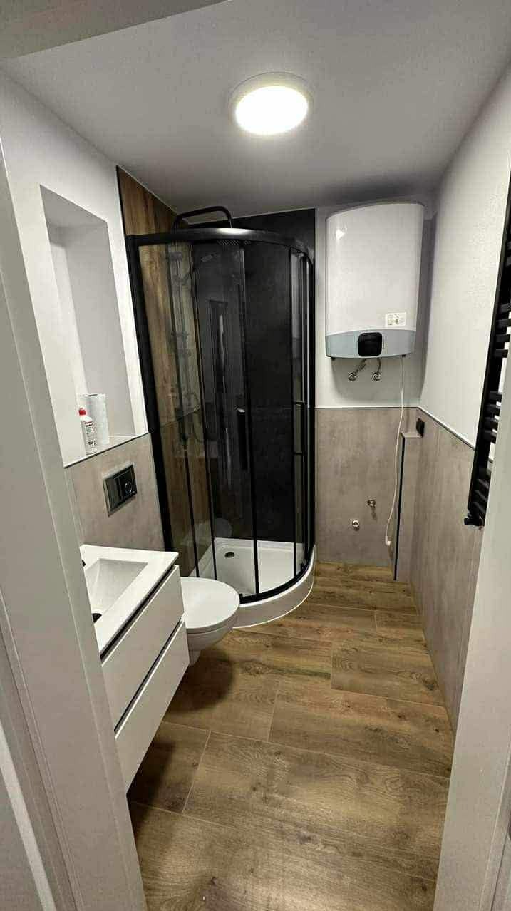 Nowoczesna łazienka po remoncie: kabina prysznicowa z czarnymi ramami, biała umywalka z szafką, drewnopodobne płytki podłogowe i bojler na ścianie.