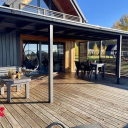 Kacper Smolarczyk - Nowoczesny dom z drewnianym tarasem i zadaszeniem w kolorze grafitowym. Na tarasie stół z krzesłami, huśtawaka i stolik kawowy. W tle zielony ogród.