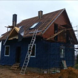 Kacper Smolarczyk - Budowa domu w trakcie: widoczne rusztowania, drabina oparta o ścianę, częściowo wykończona elewacja i dach z oknami. Na pierwszym planie zaparkowany biały bus.