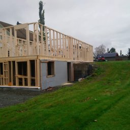 Kacper Smolarczyk - Surowa konstrukcja domu szkieletowego z widocznym fundamentem i drewnianą ramą, na tle zielonego wzgórza i pochmurnego nieba. Etap budowy z bliska.