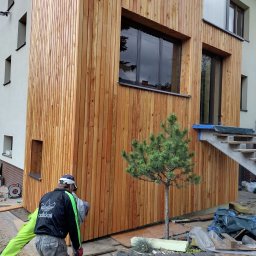 Kacper Smolarczyk - Montaż drewnianej elewacji domu z pionowych desek. Widoczny fragment budynku, okna, schody i pracownik w trakcie pracy. Małe drzewko przed budynkiem.