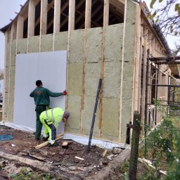 Kacper Smolarczyk - Montaż izolacji i płyty na drewnianej konstrukcji domu szkieletowego przez dwóch pracowników w odzieży roboczej. Widoczne narzędzia i materiały budowlane.