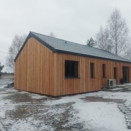 Kacper Smolarczyk - Zimowy krajobraz z drewnianym domem szkieletowym o pionowej elewacji, z widocznym klimatyzatorem i elementami konstrukcyjnymi na ośnieżonym terenie.