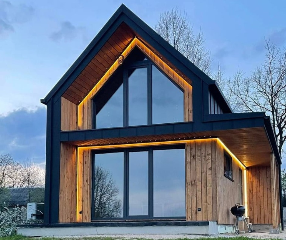 Nowoczesny dom z drewnianą elewacją i dużymi oknami, oświetlony ciepłym światłem. Minimalistyczny grill obok. Ujęcie z dołu podkreśla bryłę budynku.