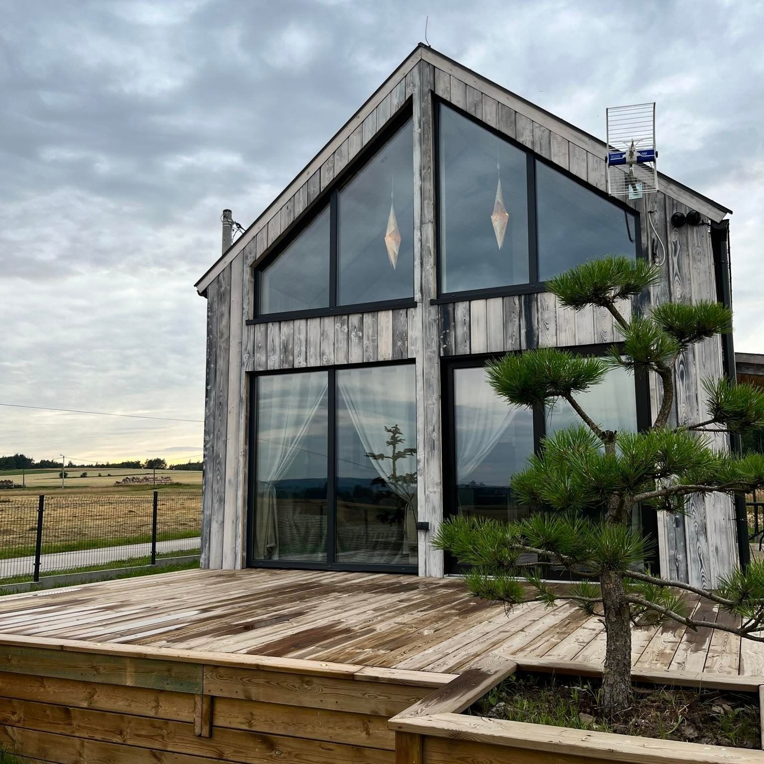 Nowoczesny dom szkieletowy z dużymi oknami i drewnianym tarasem. Widoczna antena na dachu i dekoracyjne lampy w oknach. Uwagę przykuwa małe drzewko iglaste.