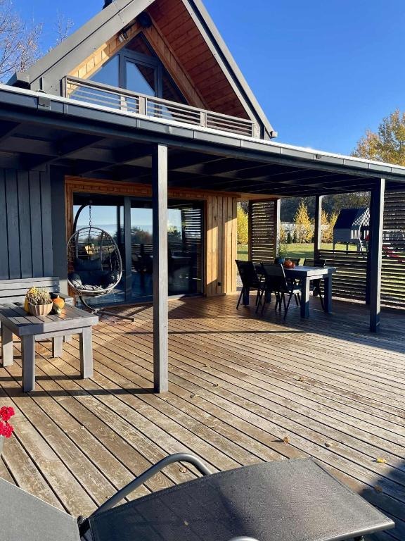 Nowoczesny dom z drewnianym tarasem i zadaszeniem w kolorze grafitowym. Na tarasie stół z krzesłami, huśtawaka i stolik kawowy. W tle zielony ogród.