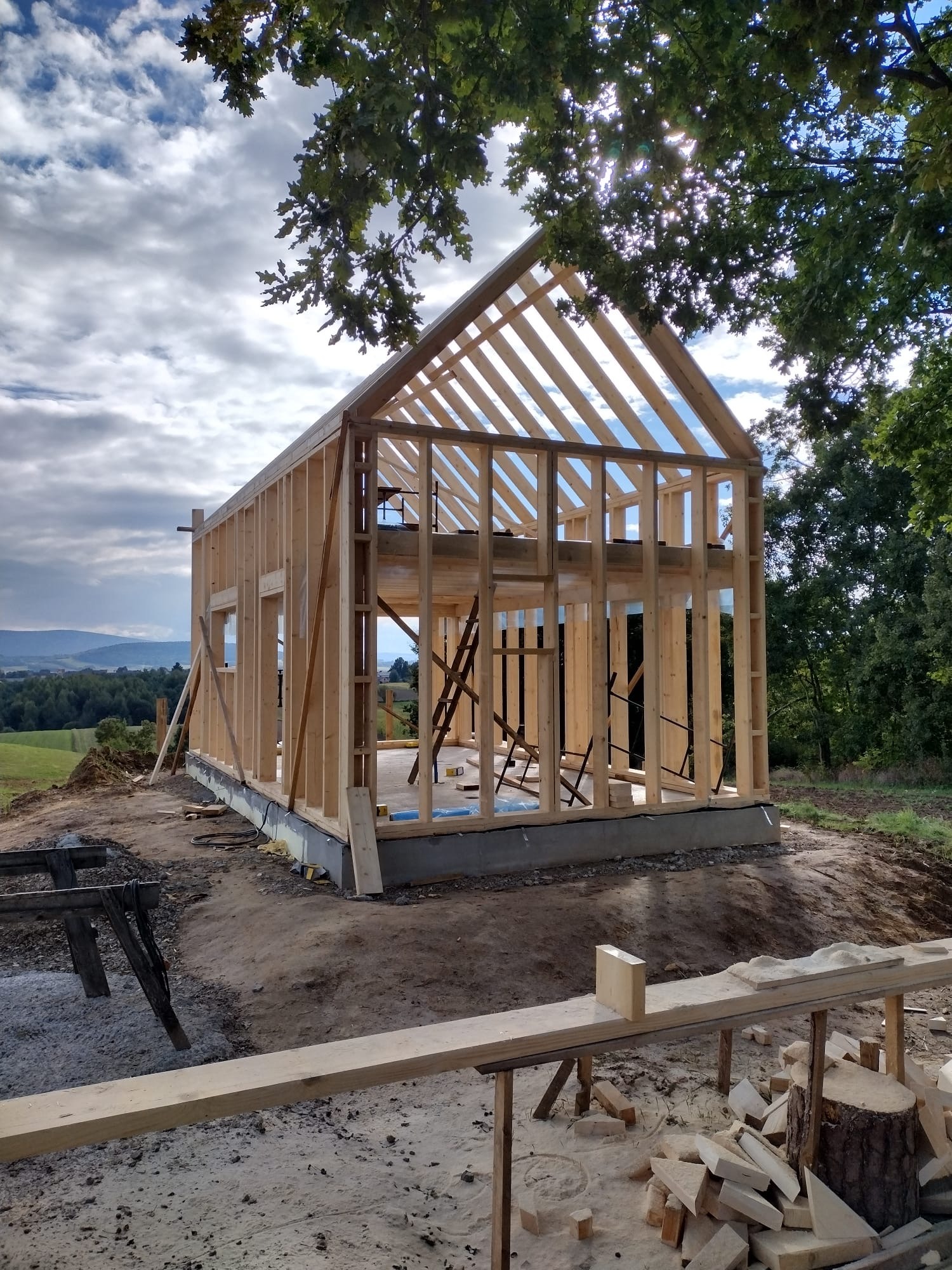 Wznoszenie konstrukcji domu z drewna na fundamencie, widok z zewnątrz. W tle zalesione wzgórza i błękitne niebo z chmurami. Częściowo widoczne drzewa.