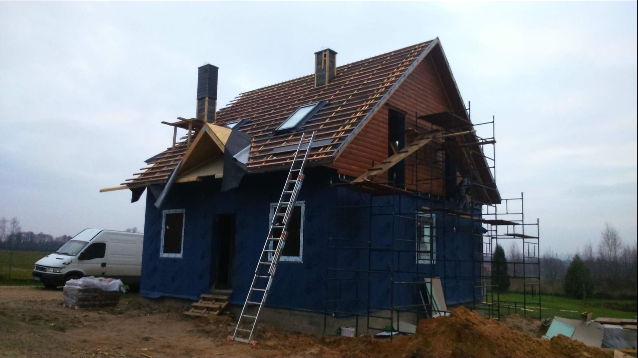 Budowa domu w trakcie: widoczne rusztowania, drabina oparta o ścianę, częściowo wykończona elewacja i dach z oknami. Na pierwszym planie zaparkowany biały bus.