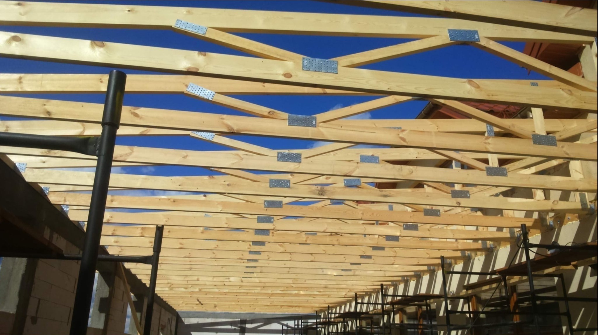Konstrukcja drewniana dachu z widocznymi łącznikami metalowymi na tle błękitnego nieba, fragment rusztowania w dolnej części kadru.