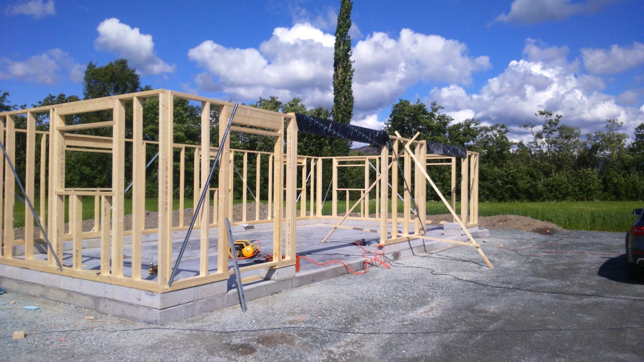 Wznoszenie konstrukcji szkieletowej domu na fundamencie. Widoczne narzędzia i elementy konstrukcyjne. Tło: zieleń, błękitne niebo z chmurami.