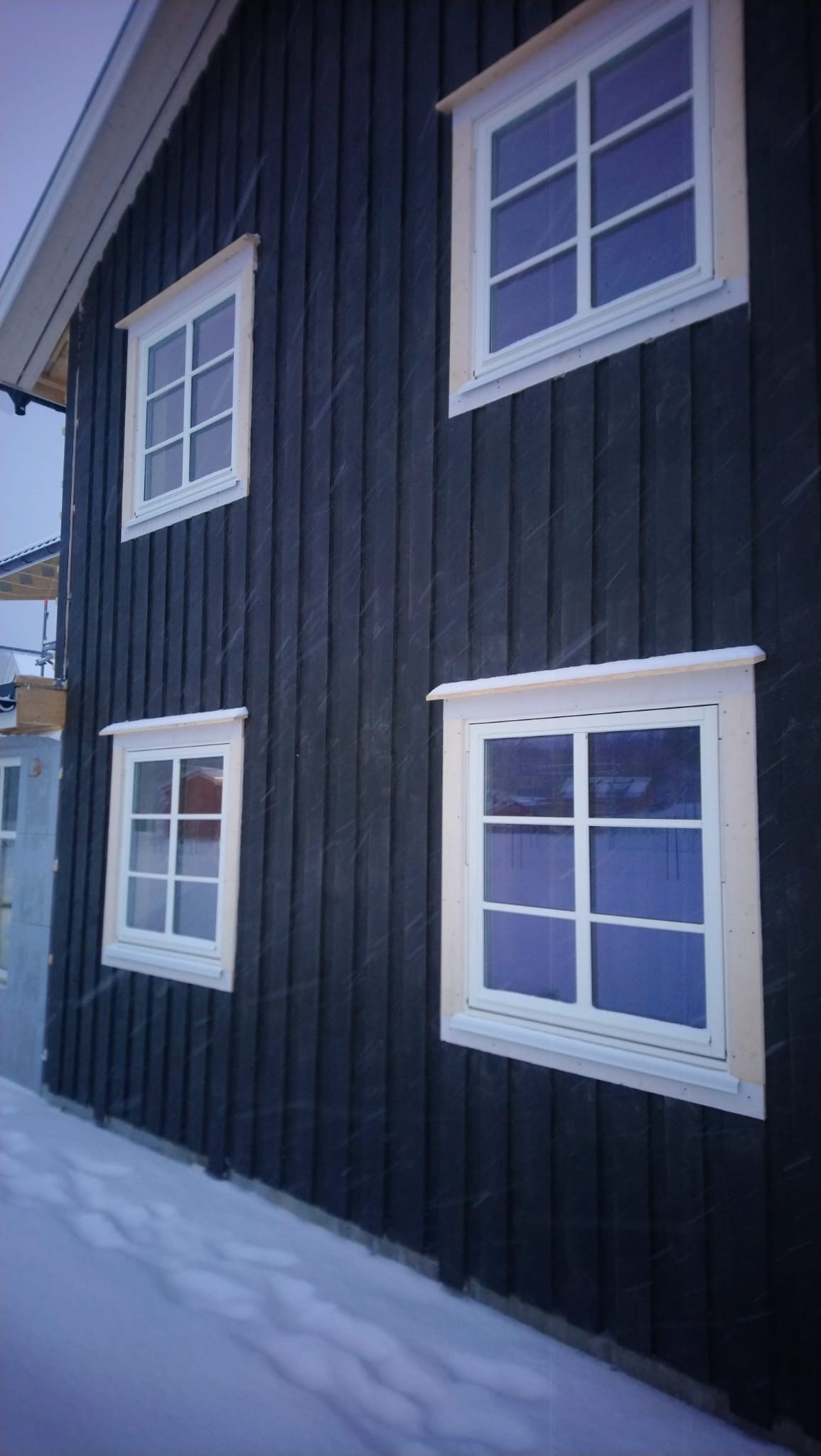 Ciemna, drewniana elewacja domu w zimowej scenerii. Widoczne białe okna z drewnianymi ramami i śnieg na ziemi. Ujęcie z dołu, podkreślające pionowe linie desek.