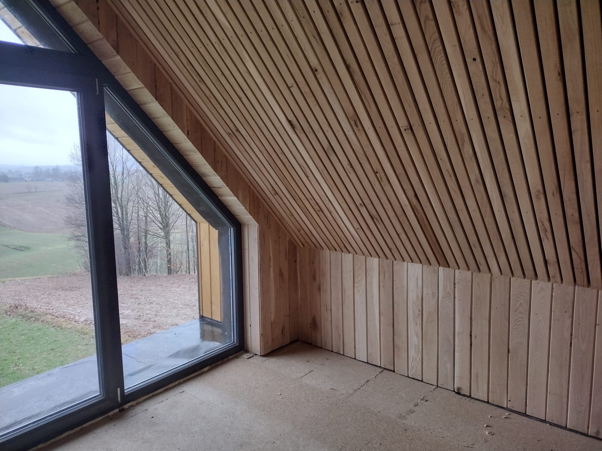 Wnętrze domu szkieletowego z drewnianą boazerią na skośnym suficie i ścianach. Duże okno z widokiem na zewnątrz. Surowy stan podłogi.