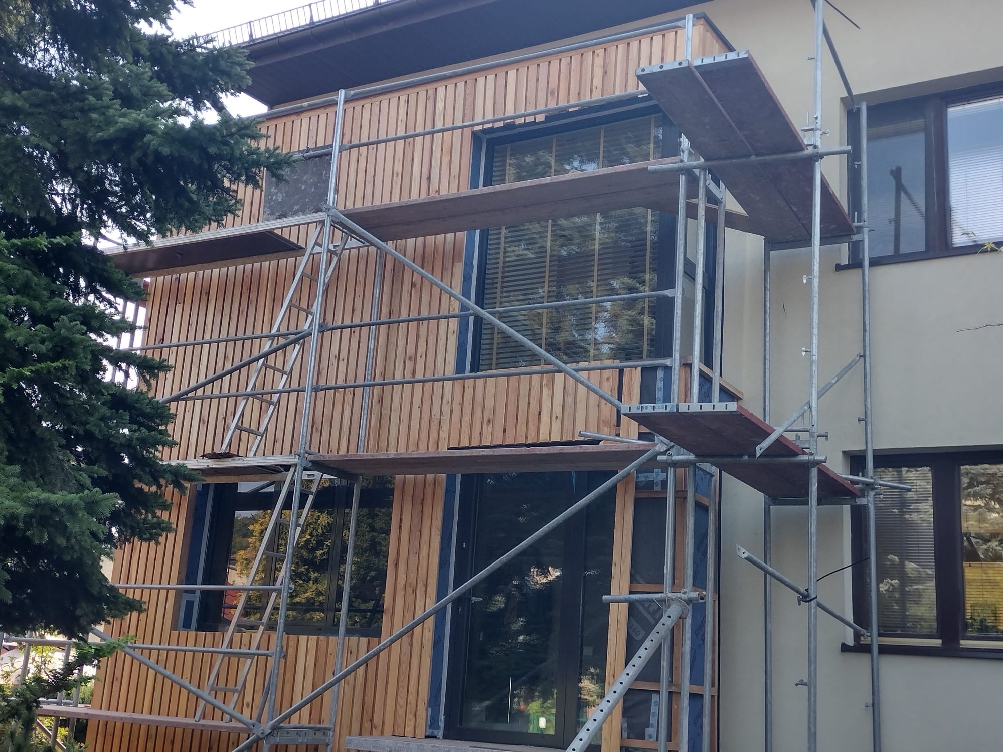 Montaż drewnianej elewacji na budynku z rusztowaniem. Widoczne deski elewacyjne, okna z żaluzjami i fragment zielonego drzewa. Ujęcie z dołu.