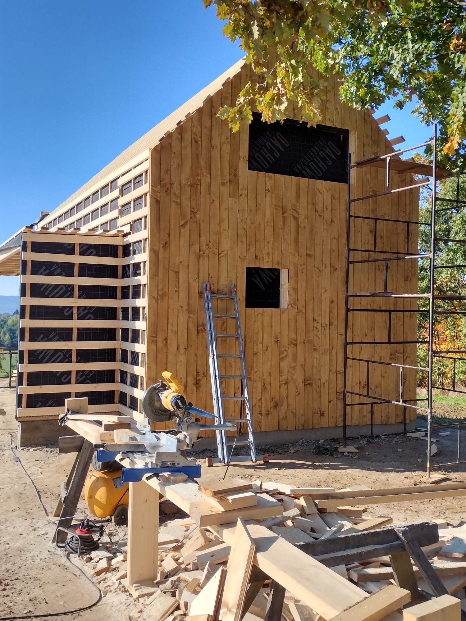 Budowa domu szkieletowego w słoneczny dzień. Widoczne drewniane ściany z izolacją, rusztowanie, drabina i piła do drewna. W tle zielone drzewa i błękitne niebo.
