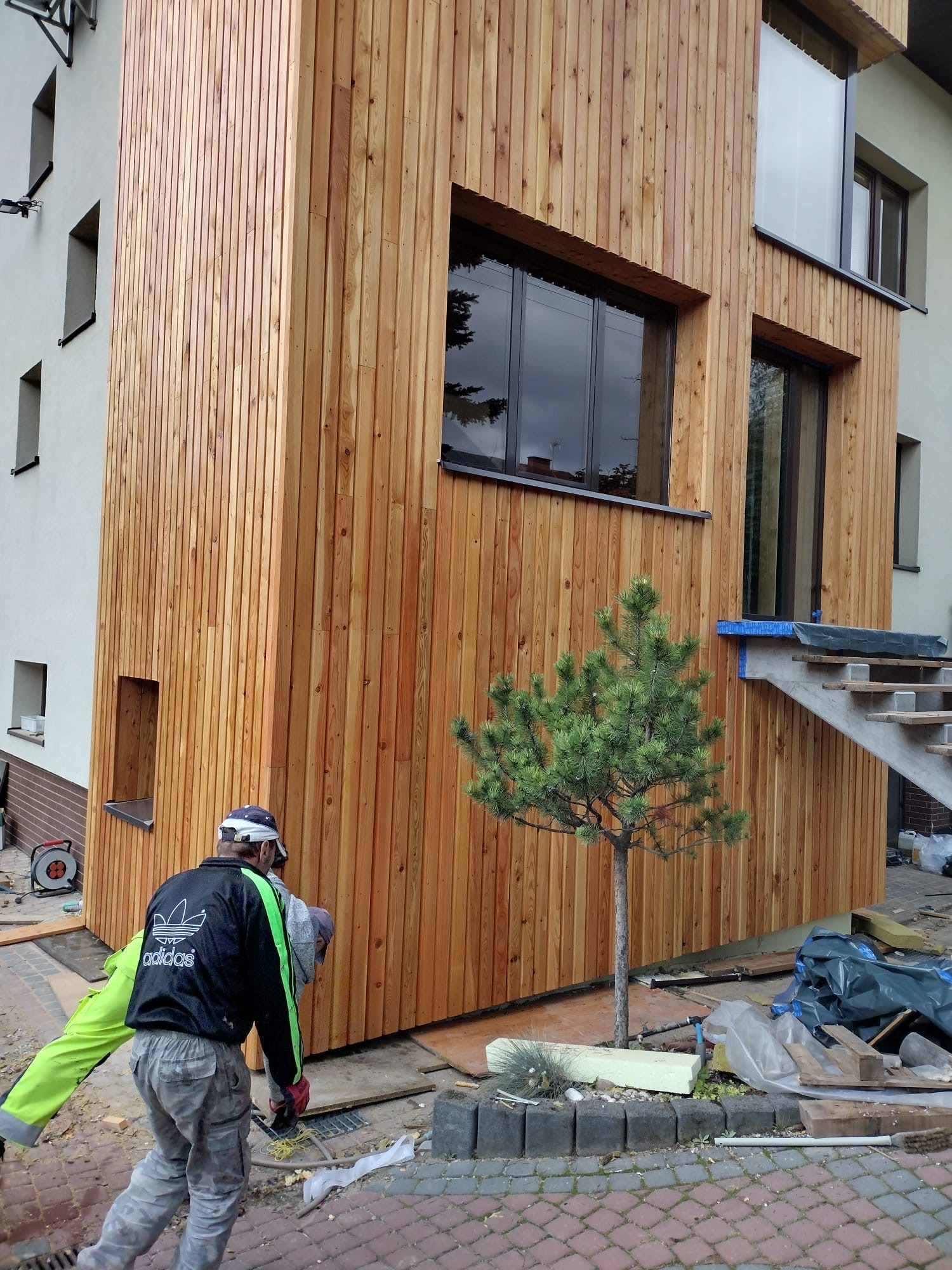 Montaż drewnianej elewacji domu z pionowych desek. Widoczny fragment budynku, okna, schody i pracownik w trakcie pracy. Małe drzewko przed budynkiem.
