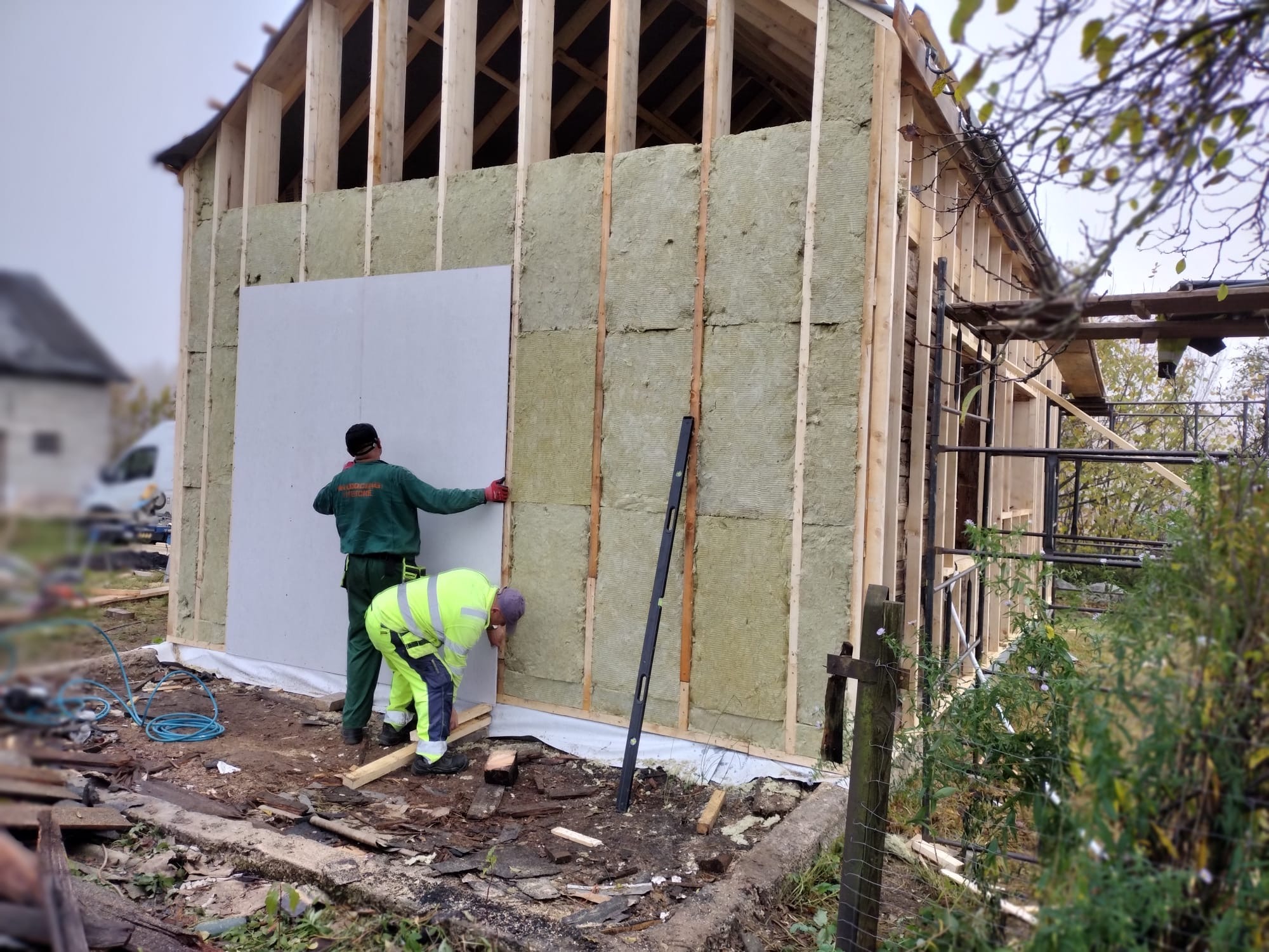 Montaż izolacji i płyty na drewnianej konstrukcji domu szkieletowego przez dwóch pracowników w odzieży roboczej. Widoczne narzędzia i materiały budowlane.