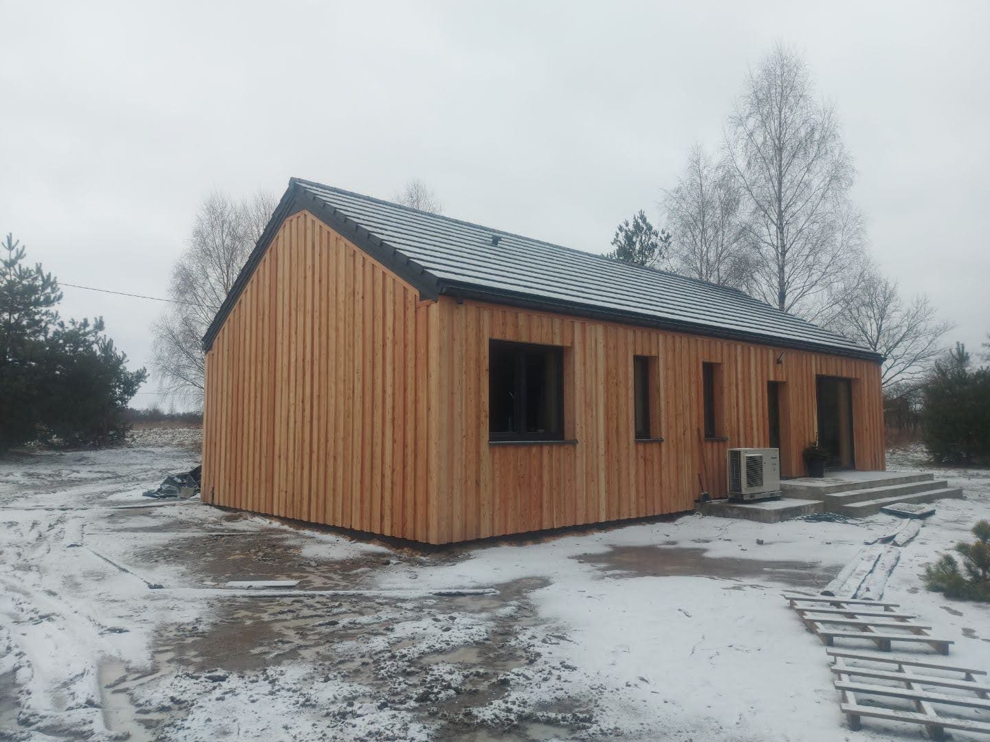 Zimowy krajobraz z drewnianym domem szkieletowym o pionowej elewacji, z widocznym klimatyzatorem i elementami konstrukcyjnymi na ośnieżonym terenie.