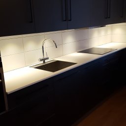 BUD-DAM - Nowoczesna, czarna zabudowa kuchenna z białym blatem i chromowaną baterią. Podświetlane płytki nad blatem. Widoczna płyta indukcyjna i drewniana podłoga.