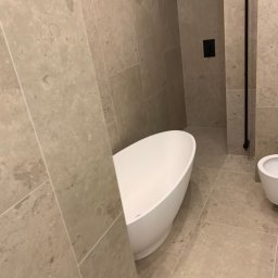 BUD-DAM - Beżowa łazienka z wolnostojącą białą wanną i bidetem. Na ścianach i podłodze płytki imitujące kamień. Czarna armatura. Ujęcie z niskiego kąta.