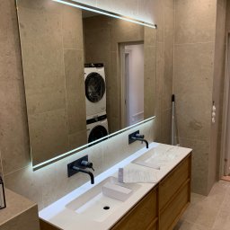 BUD-DAM - Nowoczesna łazienka z podświetlanym lustrem, drewnianą szafką pod umywalką i armaturą w kolorze czarnym. W lustrze odbija się pralka i suszarka.