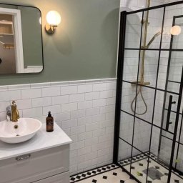 BUD-DAM - Stylowa łazienka po remoncie: kabina prysznicowa z czarnymi ramami, białe płytki, umywalka na szafce w odcieniach szarości, złota armatura i wzorzysta podłoga.