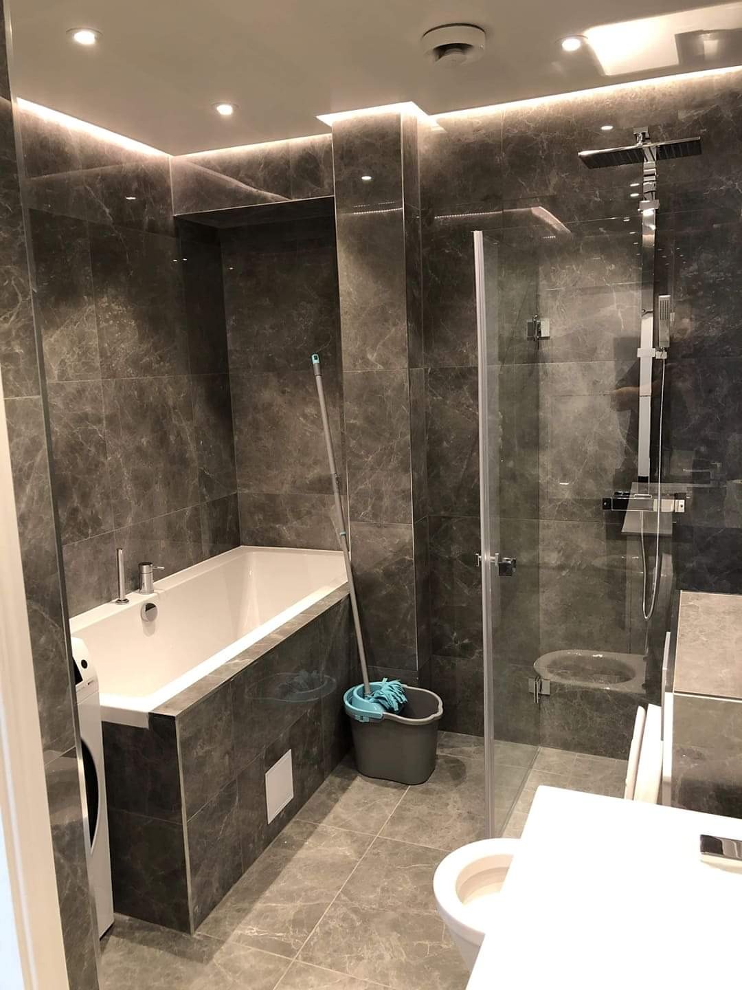 Wykończona łazienka z szarymi płytkami imitującymi marmur, białą wanną i kabiną prysznicową ze szklanymi drzwiami. Widoczny mop i wiadro. Oświetlenie LED.