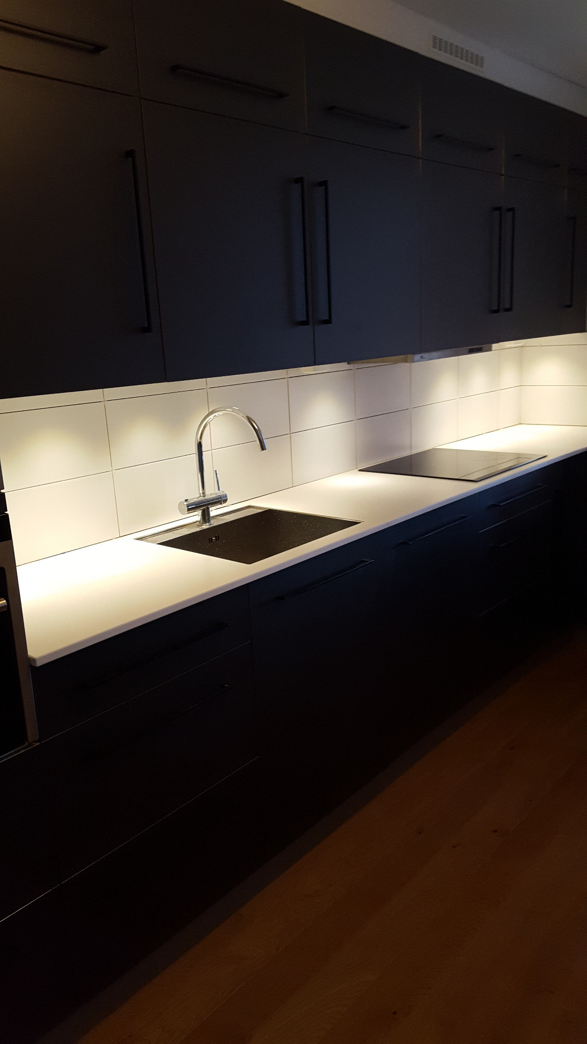 Nowoczesna, czarna zabudowa kuchenna z białym blatem i chromowaną baterią. Podświetlane płytki nad blatem. Widoczna płyta indukcyjna i drewniana podłoga.