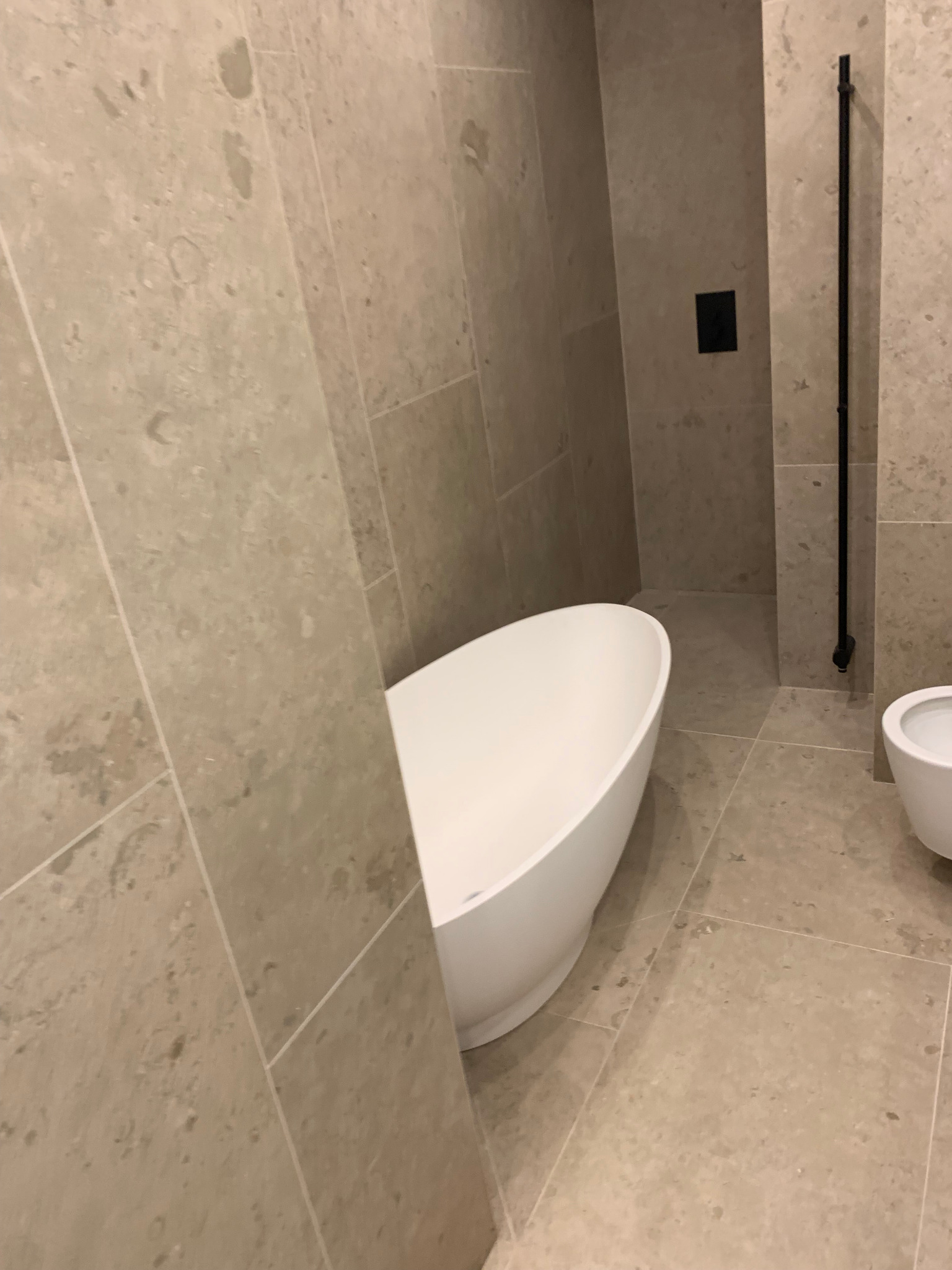 Beżowa łazienka z wolnostojącą białą wanną i bidetem. Na ścianach i podłodze płytki imitujące kamień. Czarna armatura. Ujęcie z niskiego kąta.