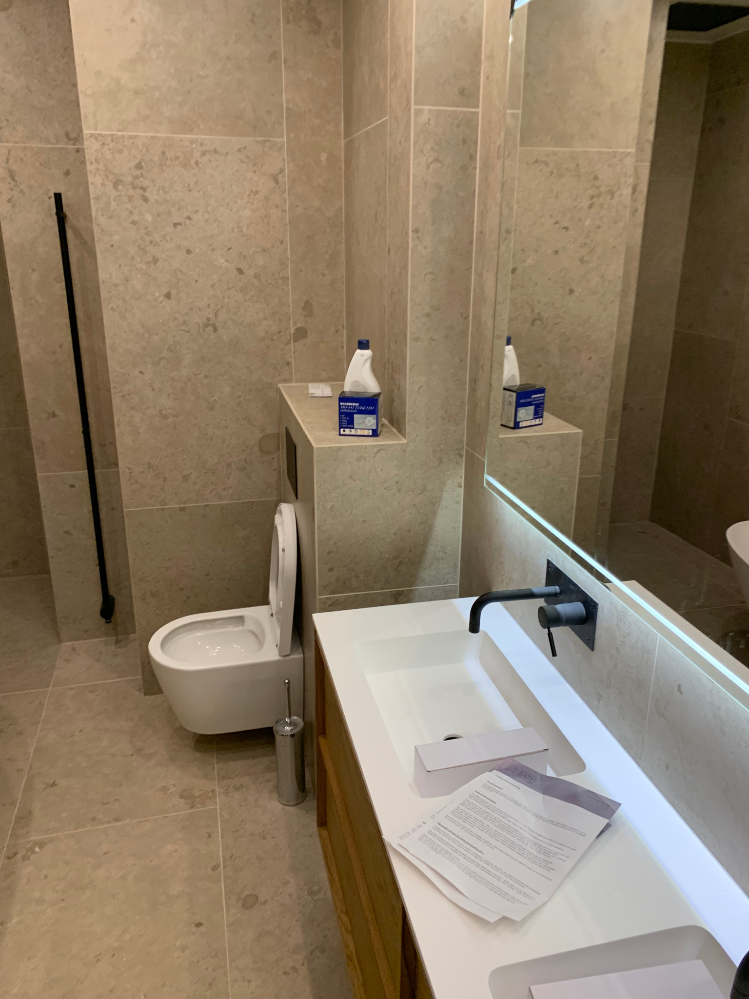 Nowoczesna łazienka z beżowymi płytkami, podwieszaną toaletą i umywalką z czarną armaturą. Lustro z podświetleniem LED. Na blacie dokumenty.