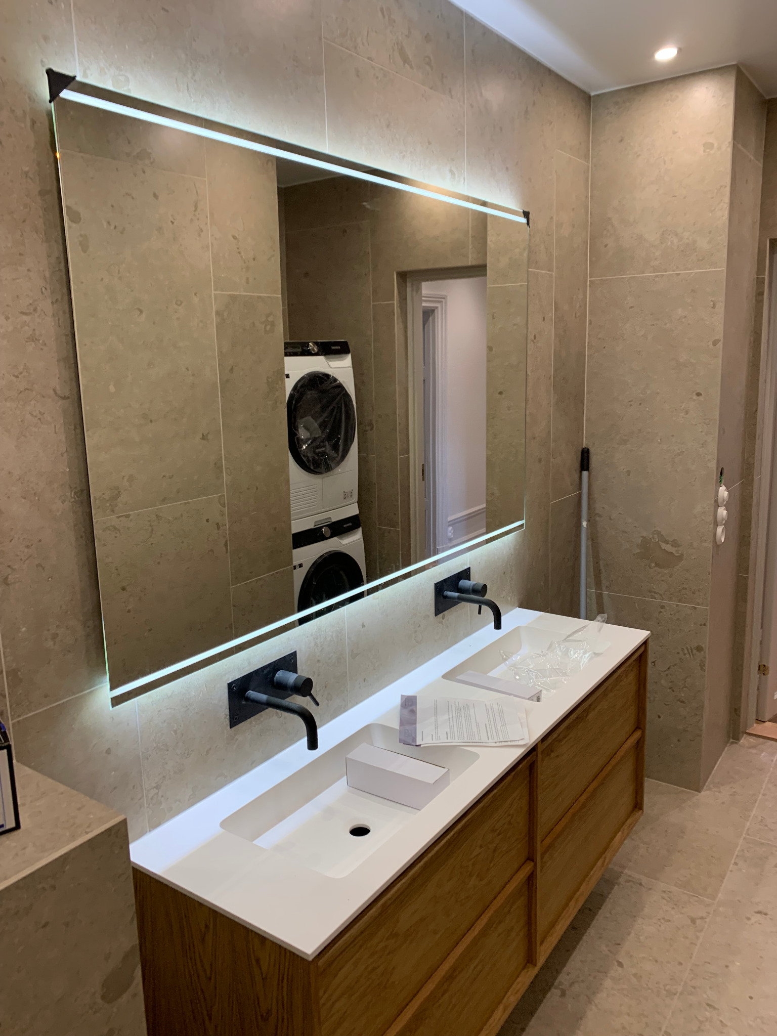 Nowoczesna łazienka z podświetlanym lustrem, drewnianą szafką pod umywalką i armaturą w kolorze czarnym. W lustrze odbija się pralka i suszarka.