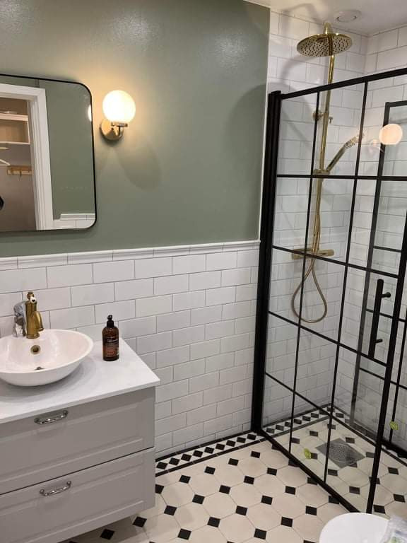 Stylowa łazienka po remoncie: kabina prysznicowa z czarnymi ramami, białe płytki, umywalka na szafce w odcieniach szarości, złota armatura i wzorzysta podłoga.