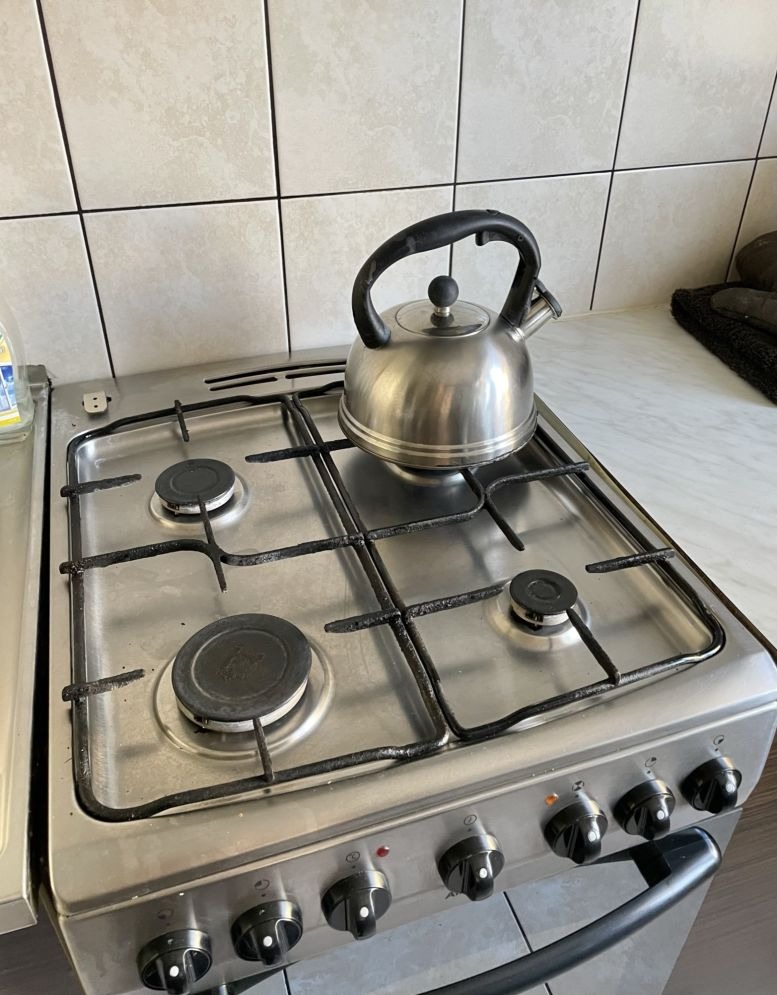 Metalowy czajnik na kuchence gazowej z czterema palnikami. Widoczne płytki na ścianie w tle. Czajnik gotowy do użycia, czeka na zaparzenie herbaty.