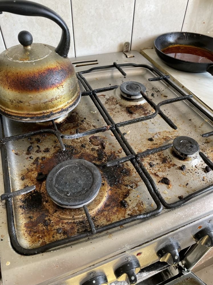 Zabrudzona płyta kuchenna ze starym czajnikiem i patelnią. Widoczne przypalone resztki jedzenia i tłuszcz. Potrzebne gruntowne czyszczenie.
