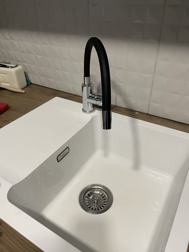 Biały, kwadratowy zlew granitowy z chromowaną baterią z czarną, elastyczną wylewką. Widoczne płytki na ścianie i fragment blatu. Czystość i minimalizm.