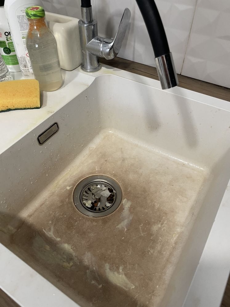 Zabrudzony, biały zlew kuchenny z resztkami jedzenia i osadem, widok z góry. Obok zlew, gąbka i butelki z detergentami. Widoczny kran z czarną wylewką.