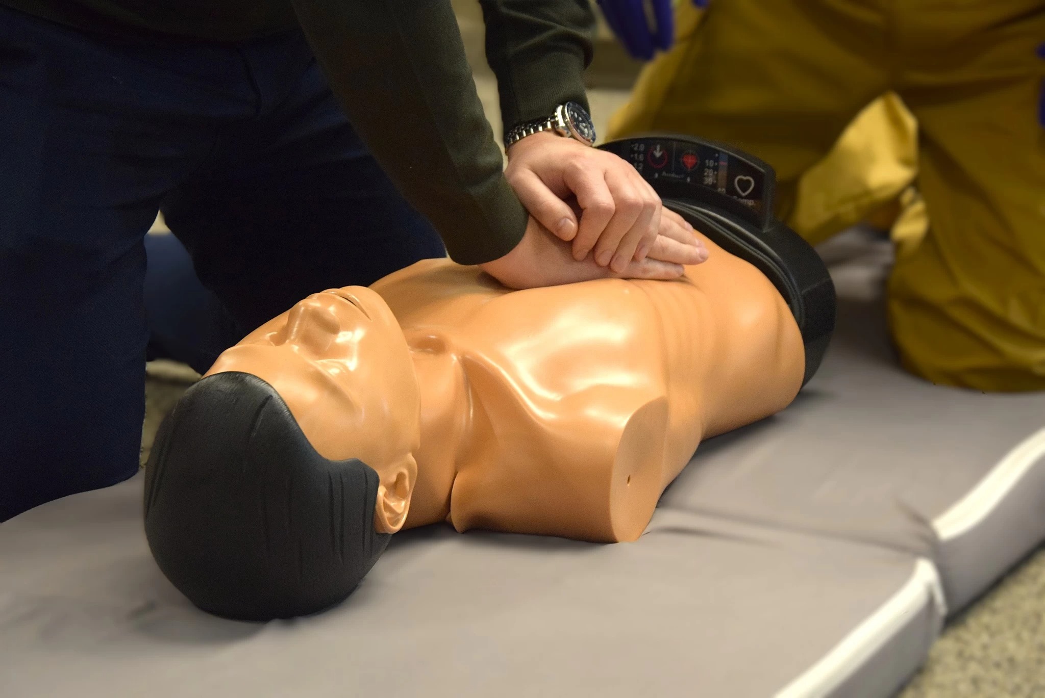 Instruktor wykonuje resuscytację krążeniowo-oddechową na manekinie podczas kursu pierwszej pomocy. Widoczne dłonie instruktora i górna część ciała manekina.