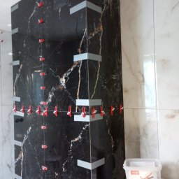 Ruslan Kotukh Usługi Remontowo-Budowlane - Czarne płytki imitujące marmur, ułożone w rogu łazienki w Katowicach, z użyciem klipsów poziomujących. Widoczny pojemnik z systemem poziomowania płytek.