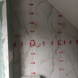 Ruslan Kotukh Usługi Remontowo-Budowlane - Układanie płytek imitujących marmur w łazience na poddaszu, z użyciem czerwonych klipsów poziomujących. Widoczny fragment toalety i surowa ściana z płyt gipsowych.