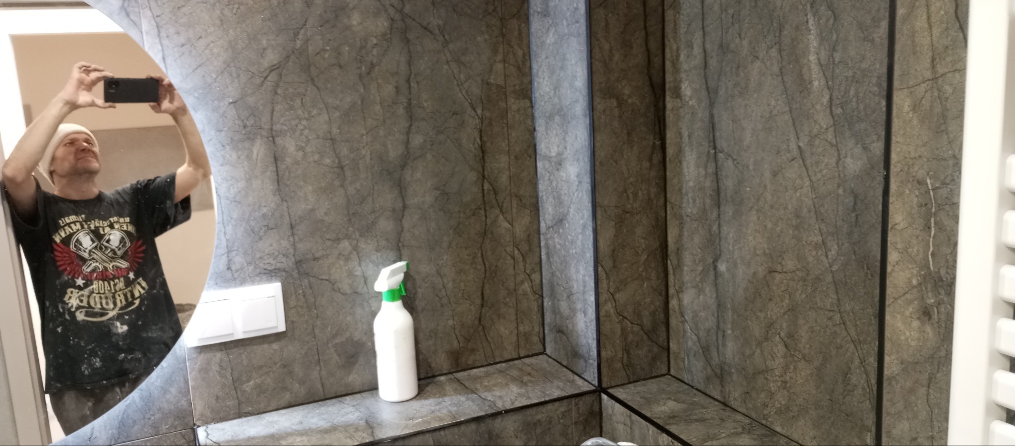 Wykończona łazienka z szarymi płytkami imitującymi marmur. W lustrze odbija się osoba robiąca zdjęcie telefonem. Na blacie biały spryskiwacz.