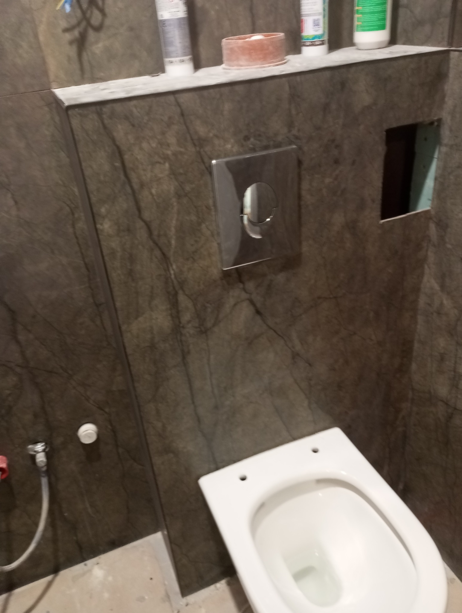 Podwieszana toaleta z szarobrązowymi płytkami imitującymi marmur. Widoczny fragment instalacji i otwór w ścianie. Minimalistyczny design, surowy klimat.