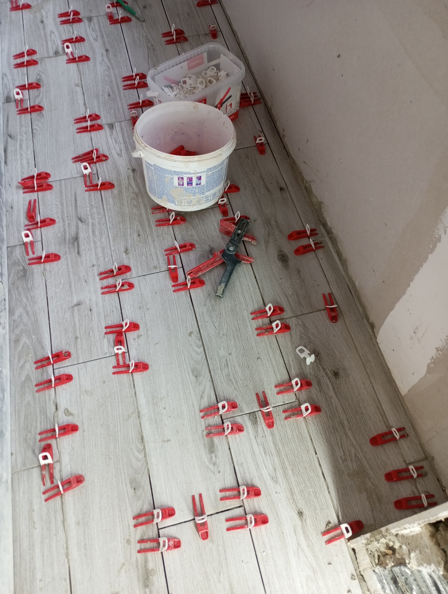 Układanie płytek podłogowych imitujących drewno z użyciem czerwonych klipsów poziomujących. Widoczne narzędzia i pojemniki z akcesoriami. Jasne, naturalne światło.