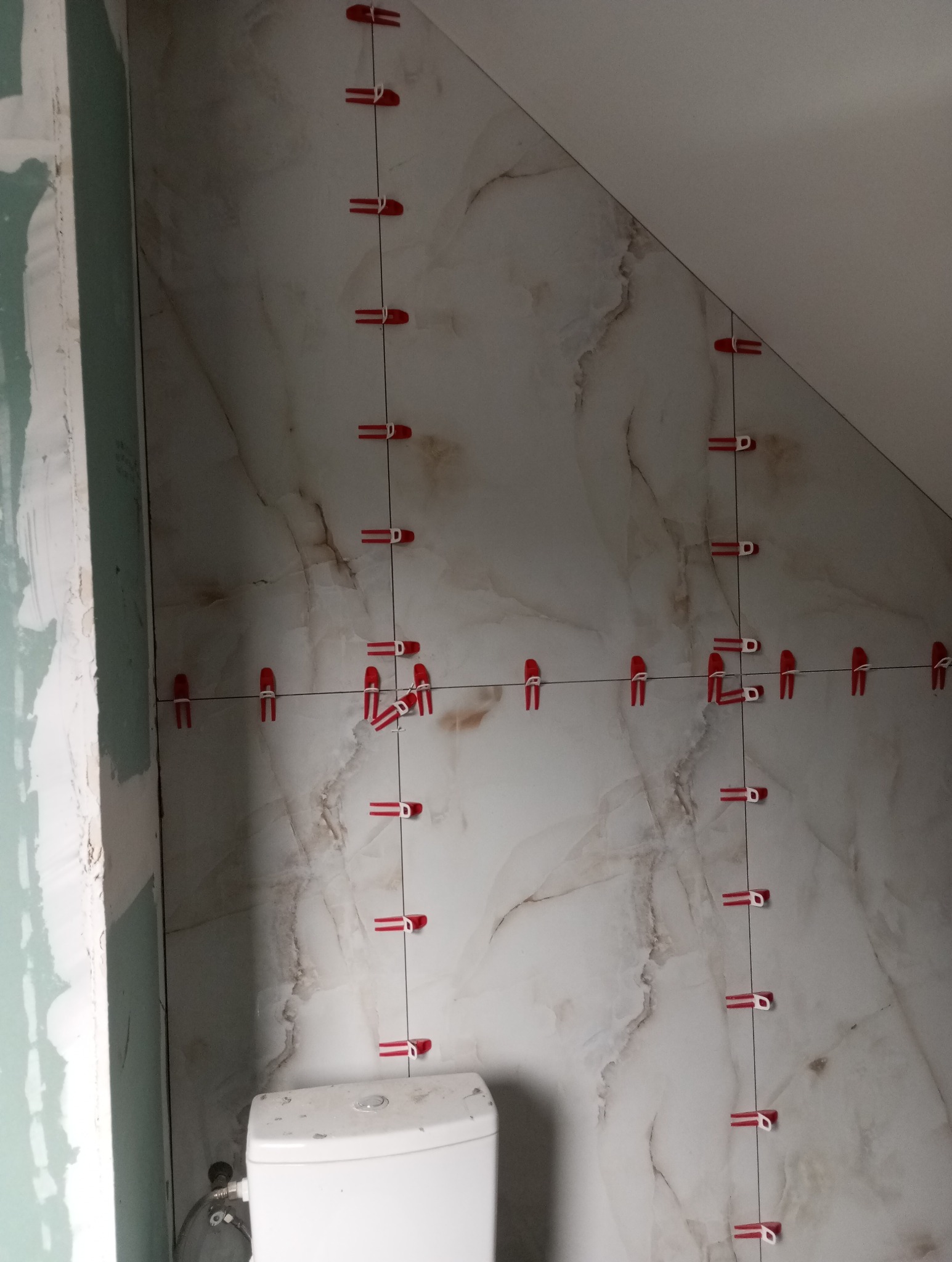 Układanie płytek imitujących marmur w łazience na poddaszu, z użyciem czerwonych klipsów poziomujących. Widoczny fragment toalety i surowa ściana z płyt gipsowych.