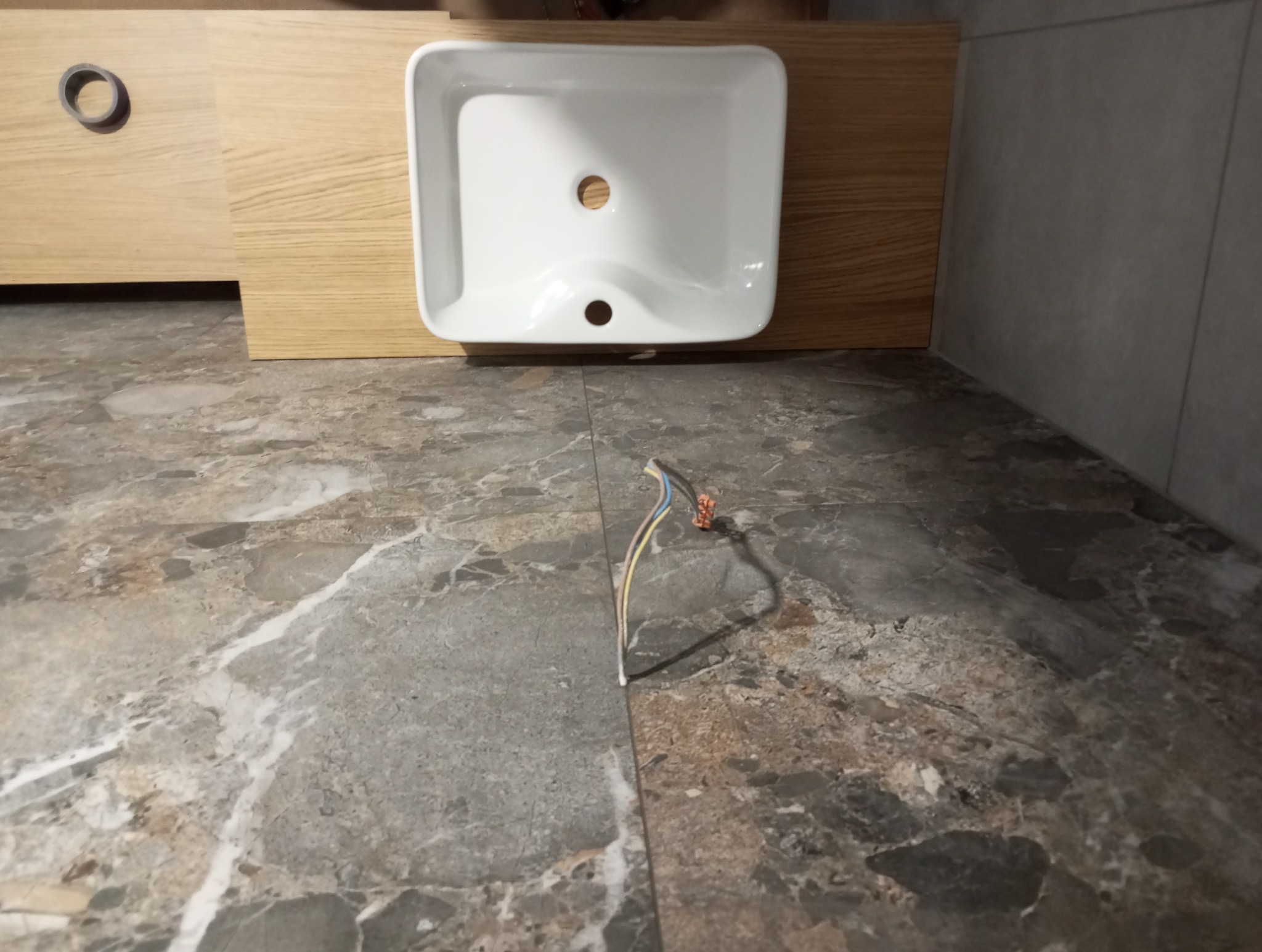 Biała umywalka nablatowa na drewnianej szafce, z widocznymi kablami na marmurowej posadzce. Ujęcie z niskiego kąta, ukazujące etap prac wykończeniowych w łazience.