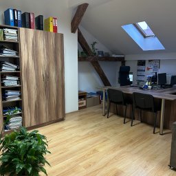 Biuro z drewnianymi meblami, regałem pełnym segregatorów i dokumentów. Na biurku komputery i kalendarze. Poddasze z oknem dachowym. W rogu doniczka z zieloną rośliną.