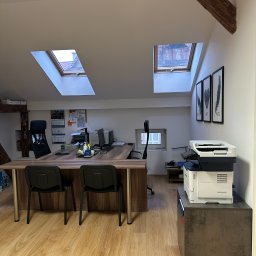 Biuro z oknami dachowymi, biurkiem, krzesłami i drukarką. Na biurku kalendarz i dokumenty. W tle widoczne ramy z grafikami na ścianie.