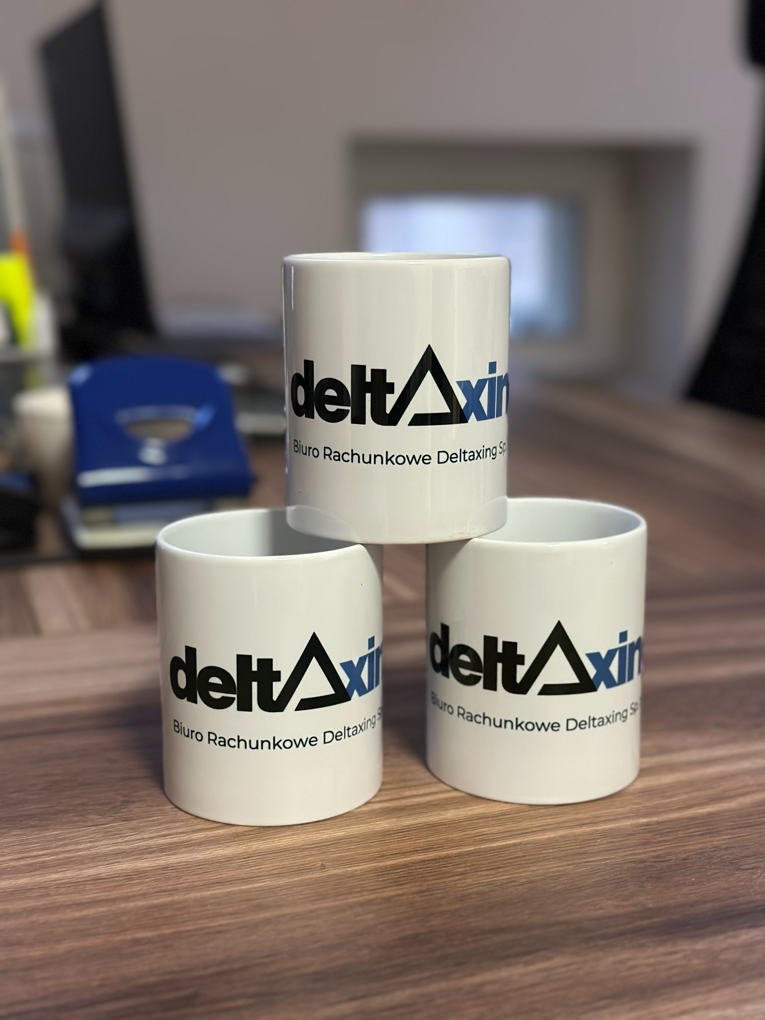 Trzy białe kubki z logo biura rachunkowego Deltaxing Sp. z o.o. ustawione jeden na drugim na drewnianym biurku. W tle biurowe akcesoria.