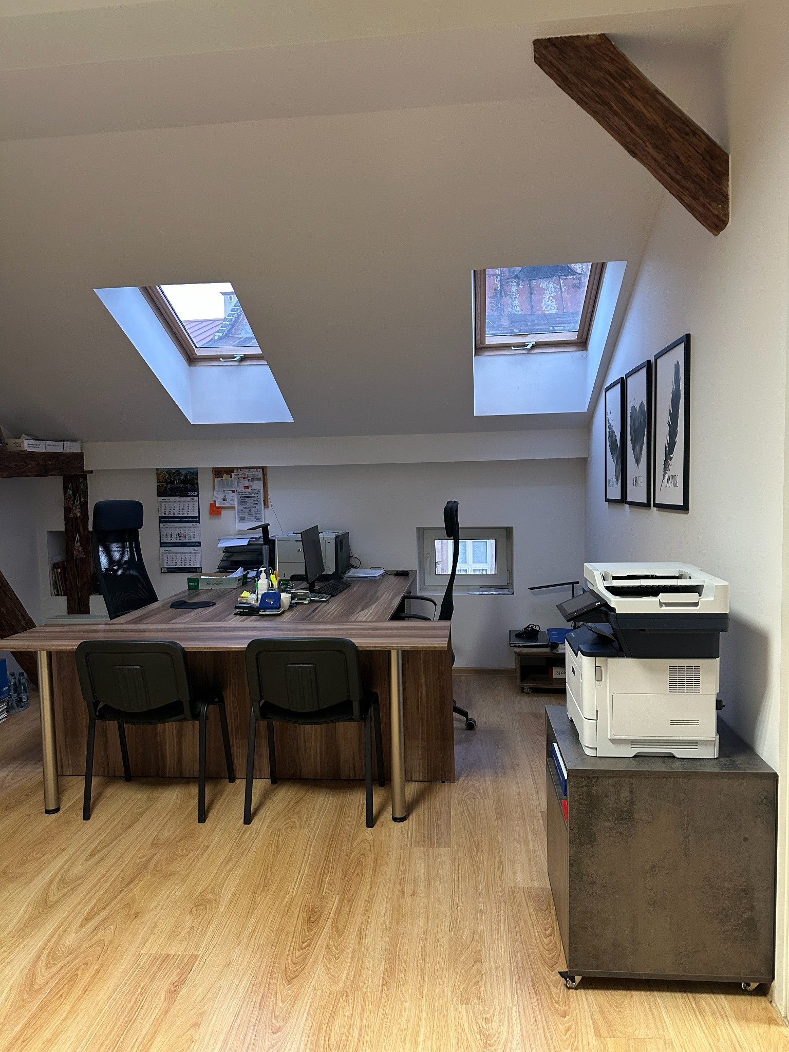 Biuro z oknami dachowymi, biurkiem, krzesłami i drukarką. Na biurku kalendarz i dokumenty. W tle widoczne ramy z grafikami na ścianie.