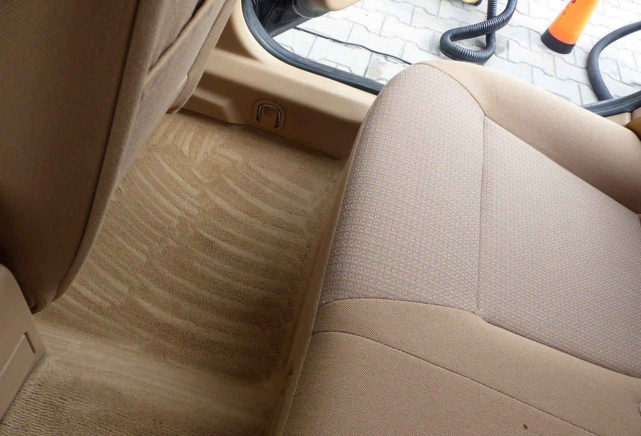 Beżowa tapicerka samochodowa po czyszczeniu, widoczne ślady po odkurzaczu na dywaniku, fotel o wyraźnej fakturze materiału. W tle wąż odkurzacza.