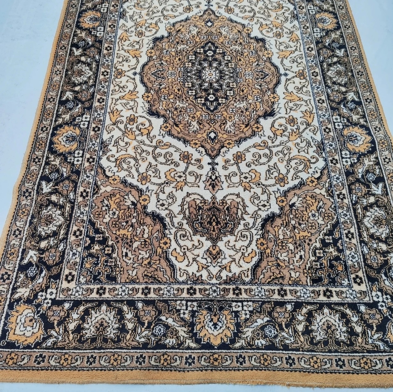 Beżowo-biały dywan z orientalnym wzorem, widoczny z góry, leżący na jasnej podłodze. Wzór dywanu zawiera motywy roślinne i geometryczne.